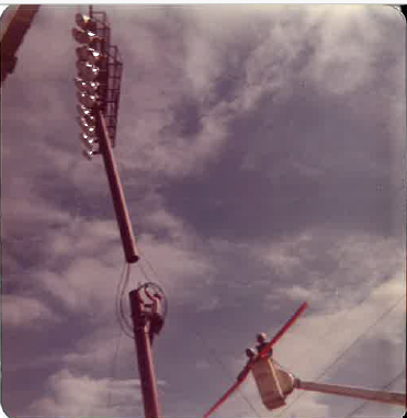 Historic photograph of a crane lifting a tall stadium lighting structure into position.