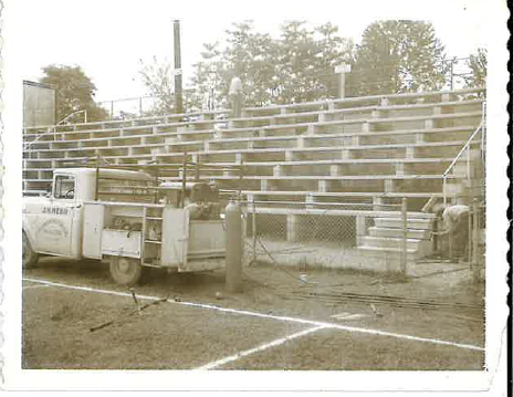 Vintage utility work truck parked beside outdoor stadium bleachers.