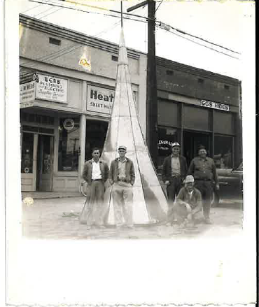 Historic photograph of five workers posing beside a large fabricated sheet-metal structure outside a plumbing and heating shop.
